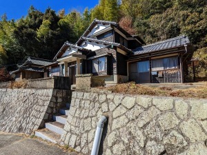 石垣の上に建っている瓦屋根、2階建ての建物で、後方が林になっている物件の写真