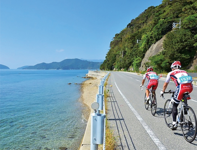 右側は山、左側には綺麗な海が広がっている海岸線の道路を2人のサイクリストが自転車に乗り、サイクリングを楽しんでいる様子の写真