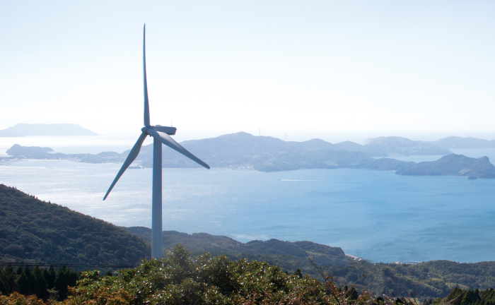 山の高所に風力発電の風車が建ち、遠くに海と山並みが見える山から見下ろした風景写真