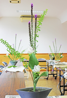 藍色の花器に細長く背の高い紫色の花と緑色の葉が飾られ、手前に白い2輪の花が活けられた生け花の作品を写した写真