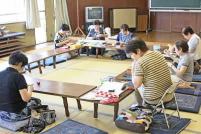 和室にロの字型に長机を並べ参加者の人達が座布団に座りながら手芸教室を楽しんでいる写真