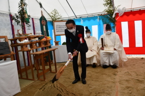 2人の神主の前でスーツ姿の男性が鍬を使って、土を掘り起こしている写真
