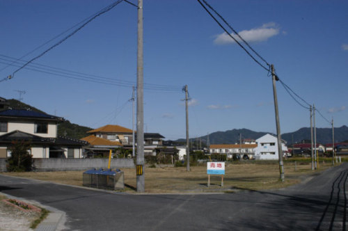 電信柱に囲まれた十字路の奥に「売地」と書かれた看板が立つ土地を写した写真