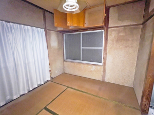 壁が一部変色しており、正面に窓、左側にカーテンがかかっている和室の写真